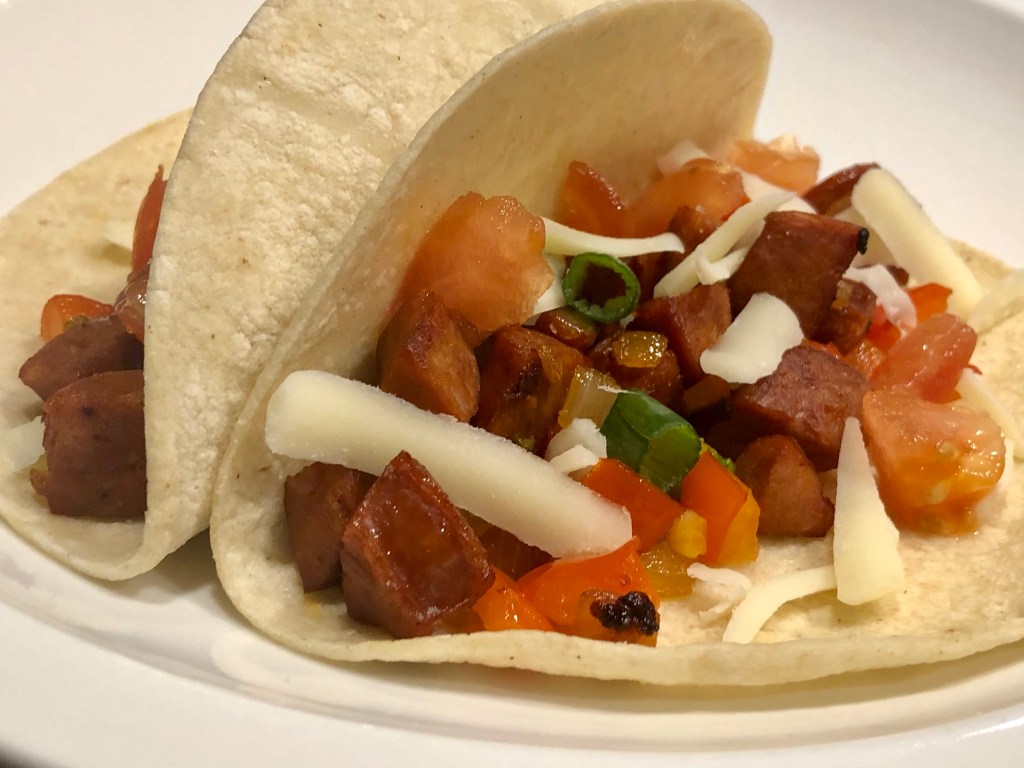 Two corn tortillas arranged as tacos; the focus is on one taco, with a mixture of diced sausage, peppers, onions, tomatoes , green onions, and sprinkled with shredded white cheese
