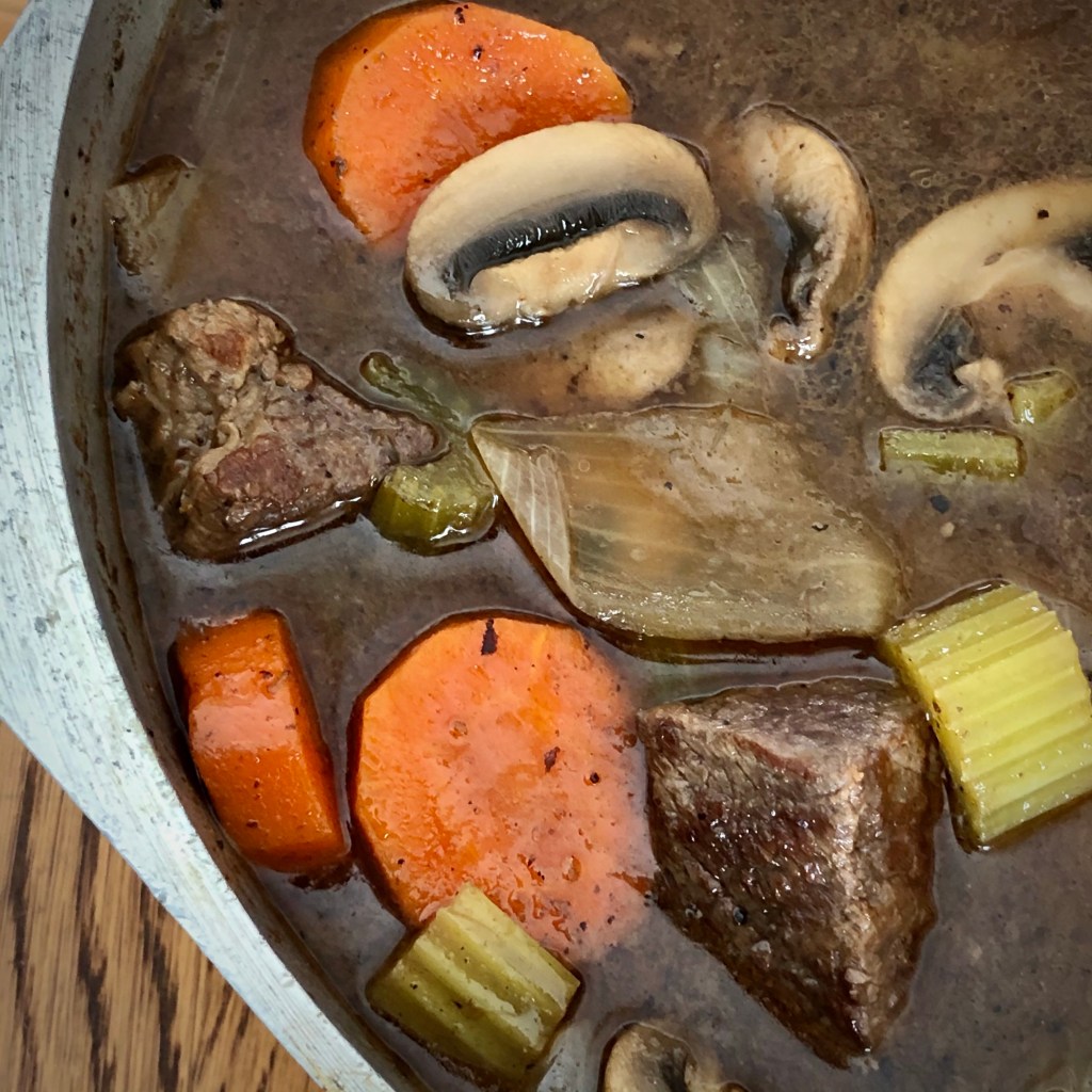 A close-up of a metal pot; inside is a stew with beef chunks, as well as slices of carrots, mushrooms, celery, and onion  